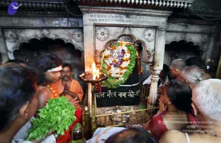 A crowded photograph inside a stone shrine niche of a temple, with a black stone deity murti dressed in a garland of white and orange marigold flowers and green leaves. A brass oil lamp with a bright flame stands in the foreground. A priest in an orange saffron shawl with a white forehead band and red tilak stands at left, holding a bundle of green leaves and a small bottle, while a man in an orange shirt with his hands folded in namaste stands beside him. Devotees press close at right, including an older woman in a red sari and several elderly men in white clothes with hands clasped in prayer. A black placard with white lettering is set against the shrine base. The scene conveys puja at the shrine.