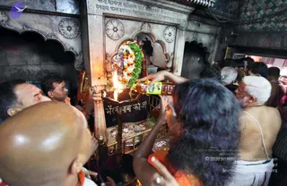 A crowded shrine interior where a woman with long dark hair in an orange top pours liquid from a plastic bottle into a brass lamp or vessel lit with a flame. The vessel sits on a pedestal beneath a stone niche garlanded with white, orange and green flowers. A shaven-headed man in white ritual cloth and a bare-chested older man with a white sacred thread stand close by, and several other devotees press in around the altar. A marble inscription in Devanagari and a black plaque reading a Bhairav name are visible on the stonework.