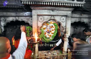 A photograph inside a stone shrine where a deity murti is decorated with white and orange marigold garlands and green leaf garlands, with a red-orange cloth draped over it. A devotee in an orange sari holds up a lit oil lamp, and a bald man in white clothing raises a hand nearby. Other people in dark vests and white shirts lean in at the carved stone railing. Black Devanagari lettering on the shrine reads 'Kal Bhairav', indicating the Kal Bhairav shrine, and the ornate grey stone carvings surround the altar.