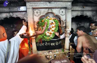 Photograph inside a dark stone shrine in Varanasi showing a black-stone deity murti decorated with a large green leaf garland, white and orange marigold garlands, and a gold-and-maroon cloth. A bald man in white clothes with an orange shawl holds up a brass aarti lamp with a lit flame toward the shrine. Another person in white reaches toward the murti, and a bare-chested man with a sacred thread stands at the right, with several devotees pressing close. Offerings and flower petals lie on the base of the shrine, and a black plaque bears the words Sri Kaal Bhairav Baba ki Jai.