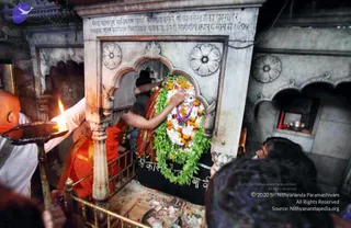 A photograph inside a stone shrine niche of a temple. A priest in saffron-orange dress reaches in to adorn a deity murti with white flowers, orange marigolds and green leaf garlands, and a purple flower necklace is visible on the murti. A white-clad attendant on the left holds a brass oil lamp with a flame toward the shrine. Carved white marble surrounds the niche, with an inscription panel above it, and devotees' heads are visible at the lower right. Scattered petals and offerings lie on the base.