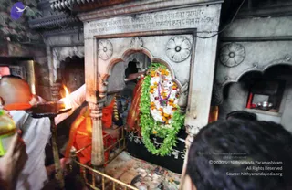 A photograph inside a stone shrine niche of a temple. A deity murti with a face and body covered in white flowers and orange marigold garlands, with a green leaf garland draped down, stands in an arched alcove. A priest in orange and red robes with a bare shaven head stands at the left holding a lit oil lamp on a tall brass stand, while another person in orange garments reaches toward the murti. Carved white marble walls with an inscription above the arch surround the shrine, and a few other devotees are partially visible at the edges, one in the right foreground. Floral offerings and a worn cloth are on the platform in front of the murti.