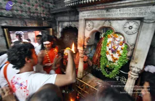 Photograph inside a dim stone shrine where a man in a white t-shirt holds up a lit oil lamp toward a deity murti heavily decorated with white and orange marigold garlands and green leaf garlands. Beside him a priest in saffron-orange dhoti with a red-and-white headband and a beaded necklace holds a brass vessel near the murti. Behind them other devotees in white and orange crowd the doorway, and ornately carved marble arches and a carved inscription frame the shrine.