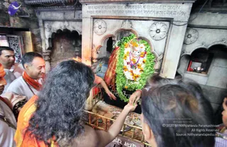 A photograph inside a stone-carved shrine niche where a deity murti is dressed in a red-and-gold garment and heavily garlanded with green leaf garlands and white and orange flower strings. In the foreground, a long-haired person in orange-saffron robes stands with arms extended toward the murti, and another person in a white kurta with an orange-red shawl and beaded mala stands beside them, smiling. Other devotees and a person in white are at the left edge and a person's head is visible at the right. A brass railing runs in front of the shrine and small offerings lie on the floor. Inscribed stone lettering is carved above the shrine arch.