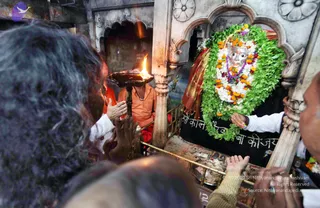 A photograph of a temple aarti at a shrine. A priest in orange robes and a rudraksha-style necklace holds a brass lamp with a burning flame toward a deity murti with a silver face, decorated with white and orange marigold garlands, green leaf garlands and a purple beaded mala. Several devotees in the foreground, including a person with grey-black hair, raise hands in prayer, and another reaches toward the altar rail. Ornate carved stone arches and pillars frame the shrine, with a black cloth draped below the murti.