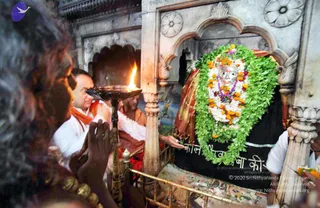 Photo inside a stone shrine in a temple niche. A priest in white clothing with an orange shawl holds a brass aarti lamp with a bright flame toward a deity murti, a silver-faced idol draped in a red and gold cloth and wrapped in a thick garland of green leaves, white flowers and orange marigolds. Behind him an attendant in saffron-orange robes stands by a brass railing. At left, a person with dark curly hair and a gold bangle reaches forward with hands joined in reverence. A second white-clad attendant is at the right edge. The black base of the murti bears carved Devanagari lettering.