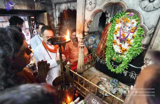 Inside a stone-carved temple shrine, a man in a white kurta and saffron-orange shawl with a rudraksha mala holds a brass oil lamp with a bright flame toward a black stone lingam decorated with green leaf garlands, white flowers and orange marigolds. The lingam bears a black cloth with white Devanagari script. A devotee in the lower left foreground has palms pressed together in prayer. A man in an orange shirt stands behind the railing, and a few other men are at the doorway. Scattered flower petals and offerings lie on a tray at the base of the shrine.