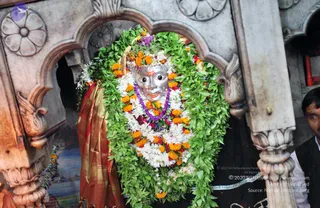 A photograph of a silver-faced deity murti decorated with a dense garland of green leaves, white flowers and orange marigolds, with a purple flower garland at its neck and a red and gold cloth draped over one side. The murti sits within an ornately carved white stone niche with lotus and floral motifs. At the right edge, a partially visible man in a dark top with a tilak stands beside the carved pillar. The scene reads as temple darshan in a shrine setting.