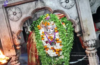 A photograph of a silver deity murti with a face and forehead tilak, decorated with a dense garland of green leaves, white flowers, orange marigolds and a purple beaded necklace, draped in a red and gold cloth. It is set within an ornate carved stone niche with lotus and floral reliefs. An oil lamp flame appears at the lower left edge. No people are visible.