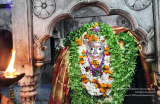 A silver deity murti with a face and red tilak sits in a carved stone niche, almost entirely covered by garlands of green leaves, white jasmine-like flowers, orange marigolds and a purple beaded garland. A gold-and-red silk cloth drapes over its shoulders. On the left, a lit oil lamp flame burns in the foreground against carved stone pillars with floral and lotus motifs. No people are visible.