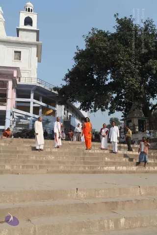 Outdoor photo of people standing on wide stone ghat steps beside a white multi-storey building with a small domed tower and a large leafy tree at right. A central figure in a flowing orange-saffron garment and orange shawl, with a rudraksha-style bead necklace, stands with arms slightly raised. To the left, a man in a white kurta and a man in white dhoti with an orange-edged shawl stand nearby. To the right, a woman in a white sari with red border and a man in a white shirt and trousers stand, and a man in a yellow shirt stands behind. A man in a checked lungi carries a bundle on his head descending the steps at right. A seated figure in orange sits at far left. Clear blue sky above.