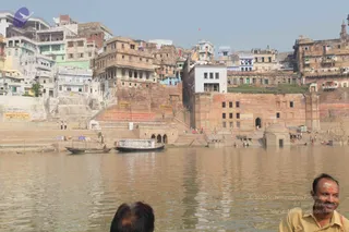 Photograph of the Varanasi riverfront across the Ganges, showing tiered stone ghats climbing to dense multi-storey buildings in faded red, tan and white. Two wooden boats sit at the water's edge near a small domed shrine. In the foreground, two men are partly visible at the bottom of the frame; the man at right, in a yellow checked shirt with a tilak-marked forehead, is the only one fully shown. Small figures are scattered on the steps and some people bathe in the water. Signs on the buildings include a shop board reading Benarasi Silk Factory.