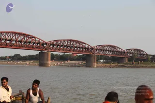 A photograph taken from a river boat looking across a wide brown river toward a long red steel truss bridge with several arched spans supported on concrete piers. Stone ghats with steps lead down to the riverbank on the left and centre, with trees and a small structure along the far shore under a pale hazy sky. In the foreground, three men sit at the edge of the boat: one on the left in a light shirt and sunglasses, one in the centre in a white sleeveless vest with a gold chain, and a partly visible bald man at the lower right wearing an orange garment. Birds are scattered in the sky.