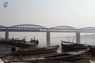 A hazy river scene on the Ganga with several wooden rowing boats moored at a ghat in the foreground, their oars and planks visible. Beyond them a long steel truss bridge with arched lattice spans crosses the river on concrete piers. A few small figures stand on the boats and the riverbank at left. Pale sky and distant hazy shoreline complete the view.