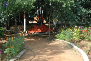 A garden path winds through shaded trees toward a seated statue draped in orange cloth, set on a raised mound of red-orange flower petals edged with dark stones. Marigolds and other flowering plants in orange, red and purple border the dirt path, with a hose running along the edge. A small sign reading PLEASE DO NOT TOUCH or similar stands at left. No living people are visible; the figure is a statue depicted in the garden setting.