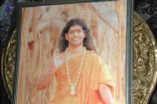 A framed printed portrait photograph set in an ornate gold-toned frame, showing a man with long dark hair, a red bindi on his forehead, and an orange-saffron shawl draped over his shoulders. He wears a long necklace of pale beads and a large pendant, and raises one hand in a blessing gesture against a soft orange background. The print is a depiction, not a person physically present, and the scene is a photograph of this printed picture.