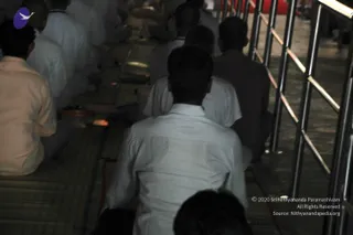 Photograph from behind of several men seated closely on mats on the floor in a dim hall, with their backs to the camera. They wear white and light-coloured shirts and trousers. A metal railing runs along the right edge of the frame. Small lit oil lamps or small flames glow on the floor near the seated devotees. The faces of the people are not visible and no deity, a person or speaker is shown.