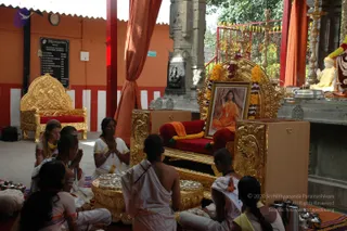 A padapuja ceremony in an open pavilion with orange walls and red-and-white pillars. A framed portrait of a smiling figure in saffron-orange robes with a long mala sits on a gilded throne draped with garlands and an orange cloth, flanked by brass vessels. Seven people sit on the floor in white dhotis and white saris; the woman centre in a white sari with yellow border holds her hands folded in prayer, while others with shaved heads hold brass plates and offerings. A second empty gilded throne stands at the left and a stone deity murti sits in a niche behind. A black board on the wall lists names beside a Dhyanapeetam sign.