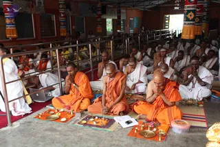 A photograph of a morning puja in a hall with orange-painted walls and colourful hanging fabric panels. In the foreground, three seated men in saffron-orange robes sit with palms pressed together in prayer before brass plates, small oil lamps and offering dishes on orange and striped mats. Behind them, rows of men in white dhotis and shawls with white forehead markings sit in prayer, and a few people in yellow and white stand along a steel railing. A laptop sits on the left near a standing figure in white.