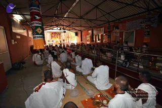 A photograph of a covered open-sided hall during a morning puja, with a row of seated devotees in white dhotis and white shawls facing a shrine at the far end. Several seated men in the foreground wear white clothing with red-bordered cloth, and some have bare shoulders; one holds a small brass plate with offerings and a small lamp sits on an orange cloth. Behind them, people stand and sit along a metal railing on the right, with oil lamps lit along the ledge. Colourful patterned pillars wrap the central area, and a deity murti is decorated in red and gold at the altar.