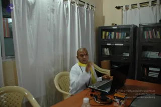 A photograph of a shaved-headed man in a white kurta with a yellow shawl draped over his shoulders, seated at a wooden table in a room with white curtains and glass-front bookcases. He wears a red tilak on his forehead and a beaded mala, and gestures with one hand while his other hand rests on an open laptop. A microphone headset, a water bottle and a small cardboard box sit on the table. Behind him, shelves hold rows of books.