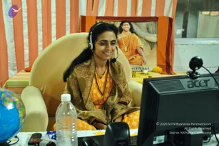 A smiling woman sits in a cream upholstered armchair at a desk, wearing a headset with a boom microphone and an orange-and-gold patterned shawl over an orange blouse, with a rudraksha-style bead necklace. In front of her is an Acer monitor with a webcam on top, a water bottle and a small globe. Behind her, orange-and-white striped drapes frame a framed poster of a saffron-robed figure with long hair and a bead garland seated before a banyan-like tree, with yellow flowers and a small lamp on a table below it. The poster's caption is partly visible. The scene reads as an online class or session setup.