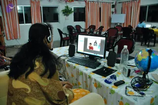 A woman seen from behind, wearing a headset with a boom microphone and a brown paisley-patterned shawl over a yellow garment, sits at a desk working at a computer monitor. The screen shows a video call with a small figure in red and another person beside them. The desk has a white floral-patterned tablecloth, a keyboard, mouse, speakers, a water bottle, a small globe and loose cables. Behind her, a room with orange-and-white striped curtains and rows of dark wooden chairs with red cushions holds a second person at another laptop. The scene looks like an online teaching or video-conferencing setup.
