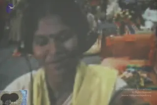 A blurred, grainy photograph of a woman in a yellow garment with a dark head covering, seated before a backdrop of flowers and an orange-cushioned seat. Her face is turned slightly toward the camera with a faint smile, and she appears to wear a headset or earpiece. The scene looks like a screen capture of video footage, with soft focus and colour blending in the background.