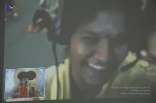 A photograph of a projection screen in a dim hall. The large screen shows a close-up of a smiling man with short dark hair, wearing a pale yellow-green top, with his face partly blurred. In the lower left corner of the screen is an inset picture of a figure in saffron-orange robes with long rudraksha malas and gold ornaments, seated before an ornate golden throne with a dark backdrop. The projected image shows a person's face and a seated figure in saffron; the room itself contains only the screen.