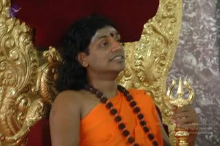 A man seated on an ornate gilded throne with red velvet upholstery speaks while holding a golden trident in his right hand. He wears an orange saffron wrap draped over one shoulder, a long string of dark rudraksha beads across his chest, and a red tilak mark on his forehead. His long dark hair falls past his shoulders. The background shows a marble-patterned wall.