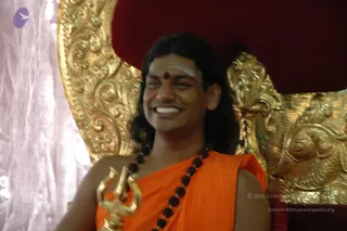 A smiling man with long dark hair sits on an ornate gold-carved throne with a red velvet backrest. He wears an orange saffron garment draped over his shoulder and a long dark rudraksha mala necklace. He holds a small golden trident-shaped staff in his hand, and a red bindi and white forehead markings are visible. A white silk drape hangs behind the throne on the left.