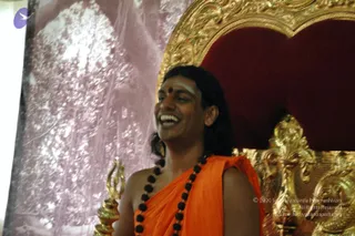 A smiling man with long dark hair and a red bindi with white forehead markings sits in front of an ornate gold and red throne with carved gilt scrollwork. He wears an orange saffron shawl over his shoulders and a long strand of dark rudraksha beads around his neck. A gold trident-like staff with a pointed finial rests beside him, and a pale pink backdrop with tree-like patterns fills the background. a person is shown in an appearance-based label, with his orange shawl, rudraksha mala and long hair as the qualifying cues, seated on the throne.