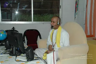 A photograph of a shaved-headed man in white ritual clothes with a yellow shawl and a rudraksha mala sits in a cream armchair speaking into a headset microphone. He gestures with one hand in front of a desktop monitor and speaker on a floral-patterned tablecloth, with a globe at left, a red chair behind, and an orange-and-white striped curtain and colourful strands at right. The setting appears to be a simple indoor room with a window showing greenery.