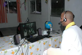 A bald man in white clothes with a yellow shawl and a rudraksha mala sits at a desk with a headset and microphone, facing a computer monitor displaying a group of people. A small world globe, a white alarm clock, a water bottle and a keyboard sit on a floral tablecloth. Orange curtains hang beside a window, and coloured paper streamers decorate the wall.
