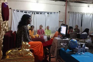A photograph of an indoor class or session in a plain room with white curtains on the back wall. In the foreground at left, a man with long dark wavy hair wears a leopard-print top over an orange-saffron dress and sits on an ornate gold-and-red throne with a gold lion-shaped armrest, speaking toward the room. A woman in a yellow-orange outfit sits nearby, and several women in pink, light green and white sit on chairs facing him. At right, a woman in an orange top and headset works at a laptop beside a computer monitor, with a blue-draped table holding a globe and water bottles. Hair and clothing colours are distinct, and the scene is evening-lit.