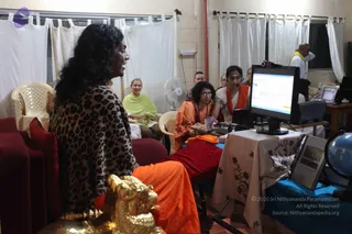 An indoor evening gathering in a plain room with white curtains and a standing fan. In the left foreground a person with long dark wavy hair and a leopard-print shawl over an orange garment sits on red cushioned seating beside a gold lion-headed carved seat. Across the room a woman in a light green wrap sits on a white plastic chair among seated listeners. At right, a woman in an orange sari wearing a headset microphone sits at a table with a computer monitor, while another woman in orange with glasses sits beside her. A globe and a small framed display sit on the table, and a man in white with a yellow shawl stands near the window.