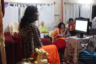 A photograph of an indoor session in a plain room with white curtains. In the foreground a woman with long dark wavy hair, wearing a leopard-print shawl over orange clothing, sits on a red-upholstered seat beside an ornate gold lion-headed armrest and speaks. Behind her an audience sits on white plastic chairs, including a woman in a lime-green shawl and others in orange and white. To the right a woman in orange and a woman in white with an orange shawl sit at a table with a computer monitor showing a slide and a small screen, draped with a floral cloth. A ceiling fan and a hanging white cloth are visible.