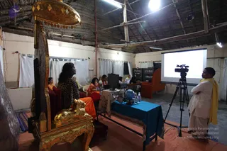 Indoor photo of a thatched-roof hall with white curtains and a projector screen showing a slide. In the foreground a large ornate gold throne with a gold canopy stands empty on the left. Beside it a woman with long dark hair and a leopard-print top sits facing the screen. Several people in orange and saffron clothing sit at tables with laptops and water bottles, and a man in white with a yellow shawl stands on the right beside a video camera on a tripod. A blue-draped table holds a globe.