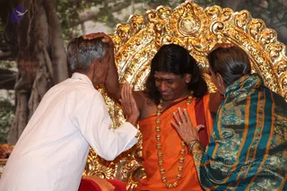Photo of a seated figure on an ornate gold throne with a carved gold backrest, with a leafy tree behind. The seated figure wears an orange saffron robe, a long gold bead necklace, and has long dark hair with a red mark on the forehead. Two people bend toward him from either side, placing their hands on his head in blessing. On the left, an older man with grey hair wears a white striped shirt. On the right, a woman wears a teal and gold embroidered sari with an orange stripe and bangles. The seated figure is smiling and looking down. a person is the seated figure: his cues are long swept-back hair, a saffron robe, a gold bead mala and a throne setting, and he is receiving blessings from the two devotees.