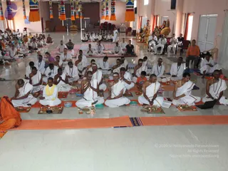 A photograph of a large hall where many men in white ritual dhotis and shawls sit cross-legged on mats in rows, facing a seated figure in orange at the left edge. In front of them are brass plates, small oil lamps, bowls and offering trays arranged on orange cloth. Several participants have white tilak marks on their foreheads and hold their hands together in prayer or sit in meditation. Hanging colourful decorative banners line the top of the hall, and a laptop sits on the floor at the right. Behind the rows, further seated devotees and a figure in white stand near a decorated throne-like seat with flowers and a golden structure. Pale pink walls and tiled floors complete the room. A pair of men in orange and white sit near the entrance on the right side. The scene suggests a group puja or satsang.