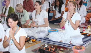 A seated group of devotees in white clothing performs a puja-style practice on striped floor mats. In the foreground a woman with dark hair in white garments and a blue cord holds her palms pressed together in prayer. Behind her a woman in white with a shaved head, a vertical forehead mark and rudraksha beads has palms joined. Beside her a woman in white with rudraksha malas and folded hands sits with eyes closed. Behind them more white-clad participants sit in meditative prayer. Brass lamps, a small flame in a dish, brass bowls and a red-striped jar sit on a rug in front of them, alongside a clay offering item. A white drape hangs in the background.