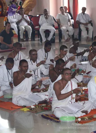 A photograph of a Pada Puja ritual in a bright hall with white marble floor. In the foreground, several shaved-headed men in white dhotis sit cross-legged on orange cloth, holding small brass oil lamps and ritual bells, with open books of chants on their laps; brass plates hold red flowers, kumkum and offerings. Their foreheads bear white vibhuti stripes and they wear rudraksha malas. Behind them, seated on chairs, men in white robes with folded hands and a man in a dark shirt watch the ritual; a golden deity figure with a cobra-hood canopy stands at the upper left. Roughly a dozen to twenty people are visible.