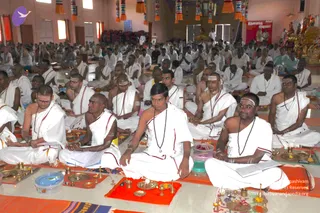 A large hall filled with seated men in white dhotis and shawls with red borders, many with white and yellow tilak marks on their foreheads and arms and rudraksha malas around their necks. In the foreground, several men sit cross-legged on orange and red mats before brass plates, cups and small oil lamps, some holding open books. Behind them rows of attendees sit on the floor, and at the upper right a decorated golden deity murti and a red banner stand near the wall. Colourful hanging tassels decorate the ceiling.