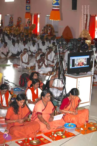 A large hall with pink walls and red curtains hosts a seated group of devotees in white ritual clothes and rudraksha malas, facing a screen. In the foreground, three women in saffron-orange saris with maroon borders sit on orange cloths, performing a puja with brass plates, small lamps, kumkum and flowers. One holds a printed sheet and another a small brass item. A tall rear monitor shows a seated figure in orange on a throne, and two cameras on tripods record the proceedings. Decorative hanging banners and deity murtis appear at the back of the hall.