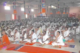 A large hall filled with hundreds of seated devotees in white dhotis and white shawls, sitting cross-legged on white mats with brass oil lamps, small plates and offering dishes in front of them. In the foreground at left, a bald man in saffron-orange robes with a beaded necklace sits facing the crowd. The hall has tall decorated pillars with orange tassels, white ceiling drapes, and a red banner with Tamil script at the back. The scene is a satsang or pada puja style gathering.
