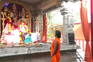 A photograph inside an open-sided temple shrine. A man in saffron-orange ochre clothing with a bare upper body stands on the right, facing the altar and looking up at the deity murti. The altar holds ornate deity figures, a seated deity with a crown and trident-like weapon, and a female figure in pink and red, all draped with garlands of purple and yellow flowers. Brass oil lamps with lit wicks line the stone platform, along with brass kalasha vessels, plates of offerings and fruit. Carved granite pillars frame the scene, with orange curtains and a red railing behind, and trees visible outside.