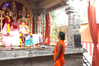 A photograph inside a stone-pillared temple shrine. A man with shoulder-length dark hair, bare-armed and wearing a saffron-orange sannyasi wrap, stands at the right facing the deity platform and looks up at the murtis. Two deity figures are dressed and garlanded with yellow and purple flower garlands; the left figure holds a trident and a crowned figure sits at centre. Brass oil lamps stand on the granite platform along with brass vessels, a plate of offerings and fruit. Red curtains hang at the open side and carved granite pillars frame the scene.