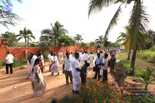 Outdoor photo of a dirt path lined by a tall red brick wall on the left and coconut palms on the right. Many people, mostly men in white shirts and white dhotis or mundus, walk and stand along the path; some women wear sarees, including a woman in a cream saree carrying a bag in the foreground. Several people wear lanyard badges. Marigold plants with orange flowers grow along the edge in the foreground.