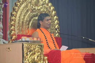 A photograph of a man seated on an ornate gold throne with a carved golden backrest and red cushions, speaking into a microphone on a stand. He wears an orange sleeveless robe over one shoulder, a long necklace of dark rudraksha beads, and has a red bindi on his forehead. He holds a small white paper in his lap. A wooden lectern in front has a gold-carved panel and a tray with fruit and sweets. The backdrop is a dark grey curtain. Appearance-based label: a person, giving a discourse while seated on a throne.