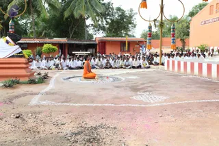 Outdoor photo of a sannyasi seated cross-legged on a raised platform in saffron-orange robes at the centre of a white kolam-decorated courtyard. Around him, a large crowd of men in white dhotis and white shirts sits on the ground in rows, many with hands folded. Behind them are orange-painted buildings, a white-and-red striped wall, tall decorative poles with ring tops and colourful tassels, and palm trees. On the left, a decorated altar with flower garlands and a white cloth sits on an orange pedestal. The saffron-clad figure at the centre is a person seated in a teaching or meditation posture, with a ritual setup before him.
